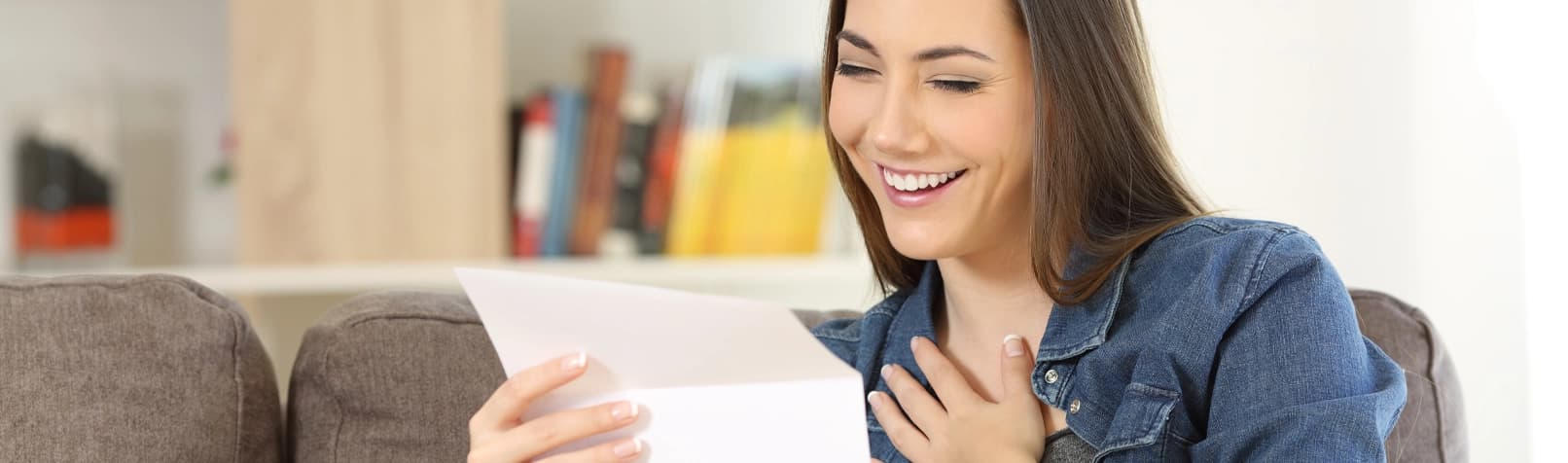 A woman smiles while opening a letter, holding one hand to her heart.